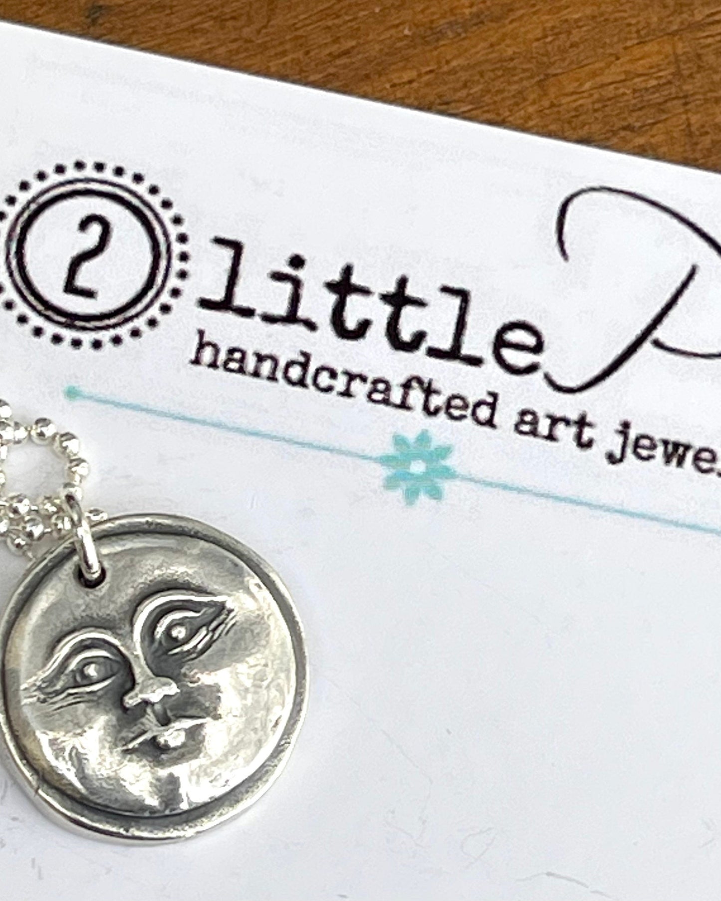 A silver necklace with a circular pendant featuring a moon face design, labeled with '2 little P's handcrafted art jewelry' on the packaging.