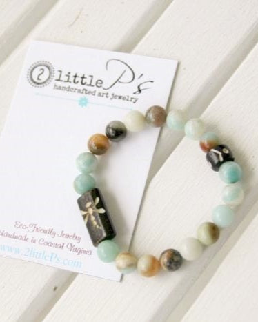 An elastic stretch bracelet with brown bone beads featuring carved dragonfly designs and amazonite tourmaline round beads, displayed on a white wooden surface with a branded card.
