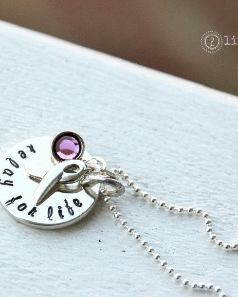 A sterling silver necklace with a large circular charm hand-stamped with 'Relay for Life,' a Swarovski crystal, and a sterling silver awareness ribbon charm, attached to a ball chain.