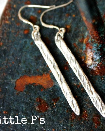 A pair of textured silver earrings laid on a rustic surface, with the brand '2 little P's' mentioned.