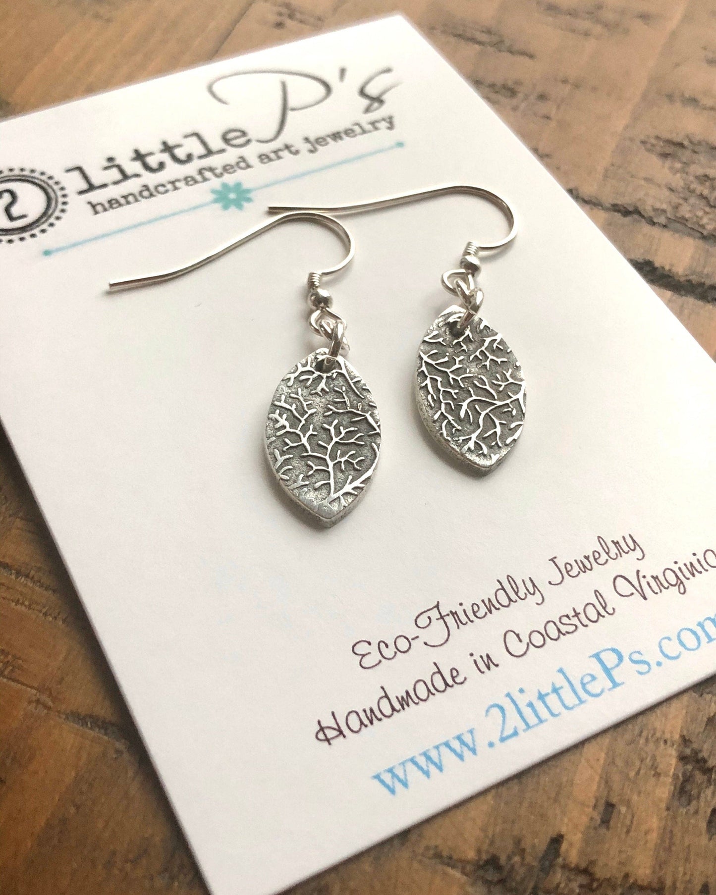 A pair of silver leaf-shaped earrings on a white card with '2 Little P's' branding and website address.