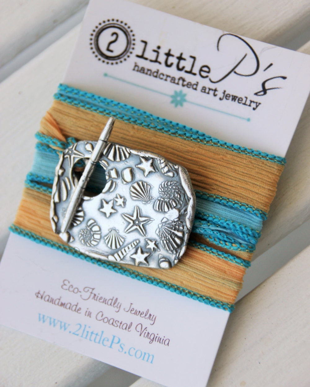 An artisan silk wrap bracelet with a pure silver toggle, featuring sea-inspired colors, presented on a card with the 2 little P's brand.