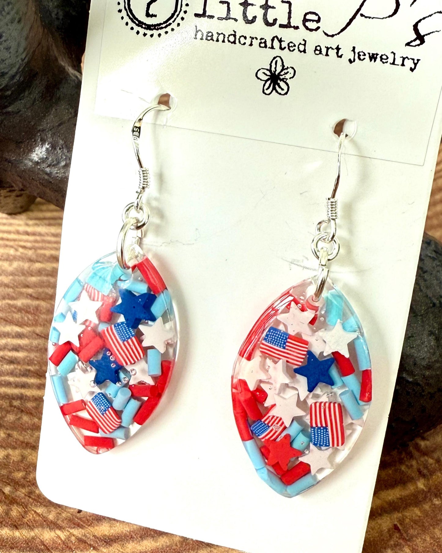 A pair of oval-shaped resin earrings with a patriotic design, featuring stars and stripes in red, white, and blue colors, attached to a card with '2 little p's' branding.