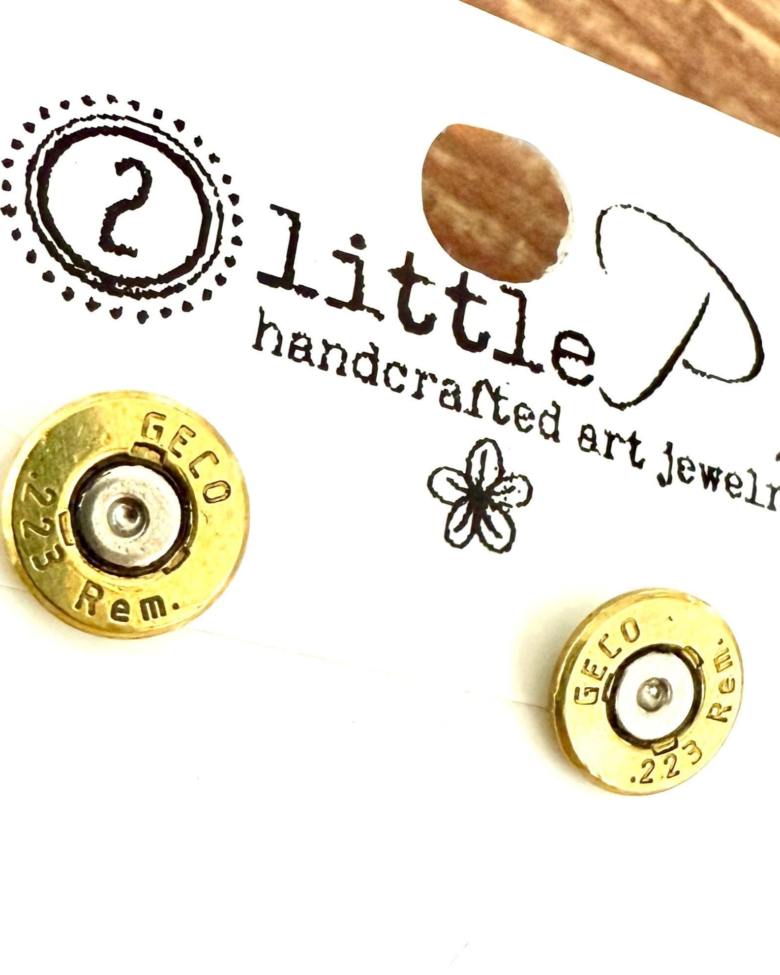 A pair of .223 Remington brass shell casing stud earrings on a branded card reading '2 little P's handcrafted art jewelry.'