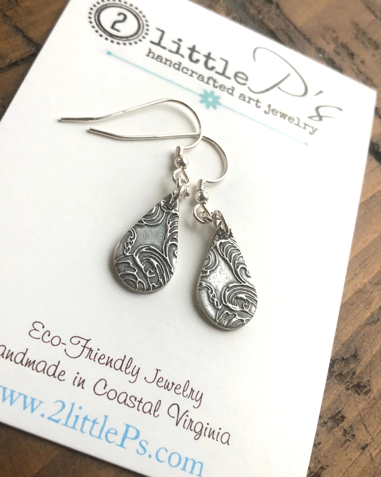 A pair of silver earrings with etched designs, presented on a card that reads '2 little P's handcrafted art jewelry'.