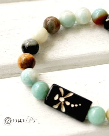 An elastic stretch bracelet with a variety of beads including brown bone beads with carved dragonfly designs, and Amazonite Tourmaline round beads in shades of blue and white, displayed on a wooden surface.