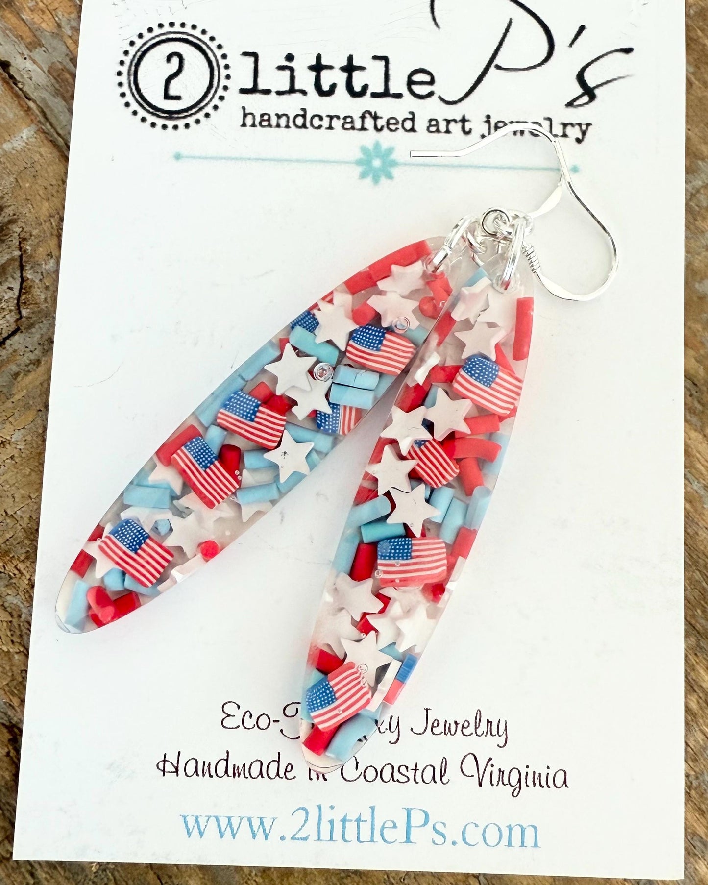 A pair of handcrafted resin earrings featuring red, white, and blue stars and American flags, displayed on a card indicating the brand and website.