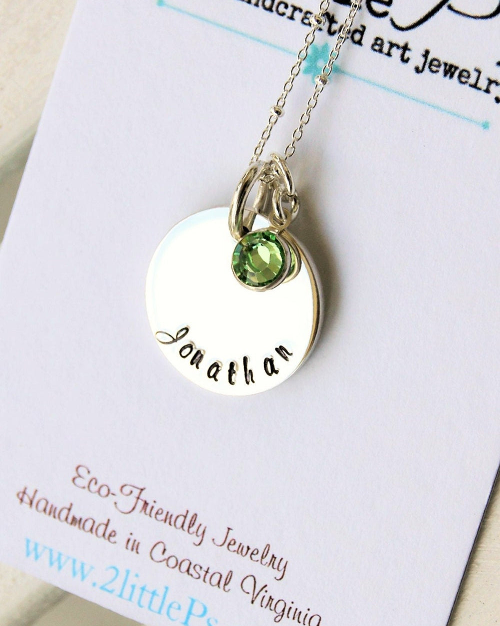 A sterling silver necklace with a hand-stamped circle charm that reads 'Jonathan' and a green Swarovski crystal birthstone drop, presented on a card with the brand name '2 little p's'.
