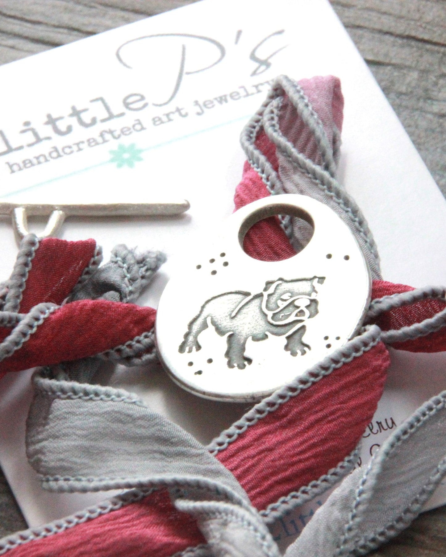 A handcrafted pure silver bulldog wrap bracelet with a handmade bulldog toggle ornament, suspended from hand-dyed shades of maroon and gray sari ribbon, displayed on a card with the 2 Little P's branding.