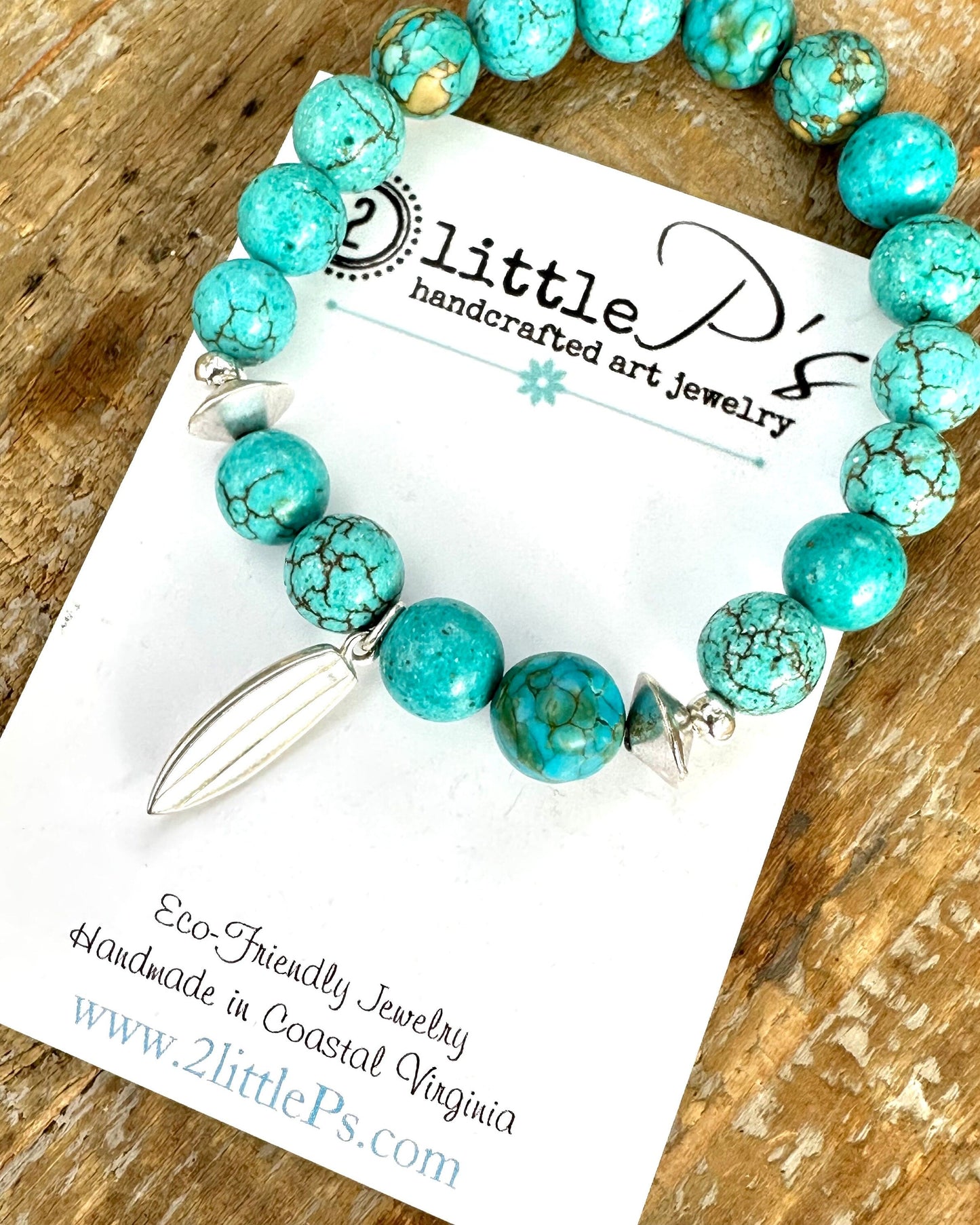 A stretch bracelet with round blue-green beads and a sterling silver surfboard charm, displayed on a card with the brand name 2 little P's.