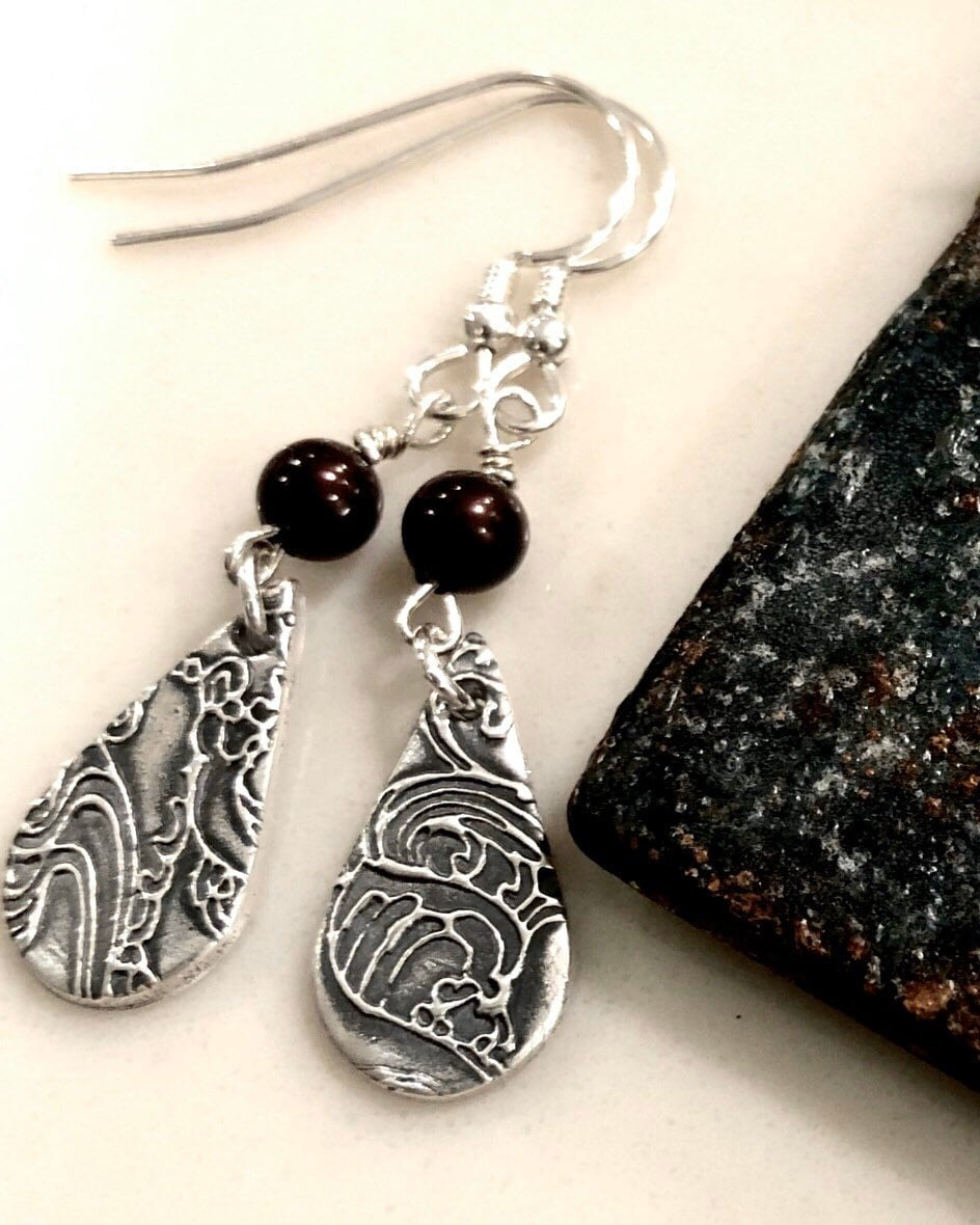 A pair of dangle earrings made of fine silver with embossed wave-like patterns and garnet beads, displayed against a stone background.